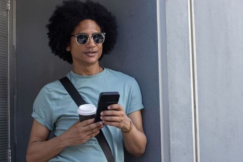 African american man leaning on wall wearing sunglasses checking phone while holding coffee cup