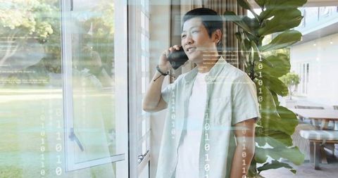 Midadult asian man talking on smartphone by window with binary overlay remote work tech