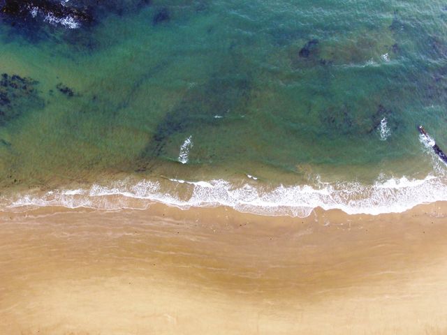 Aerial view showing clear, calm ocean meeting sandy beach below. Green waters gently touching sandy shore creating peaceful, relaxing atmosphere. Ideal for travel brochures, website backgrounds, relaxation-themed promotions, and nature photography portfolios.