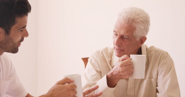Senior man enjoying conversation with a caregiver while holding coffee cups. Depicts themes of caregiving, healthcare, elderly support, and companionship. Useful for content related to aging, senior care services, and family support.