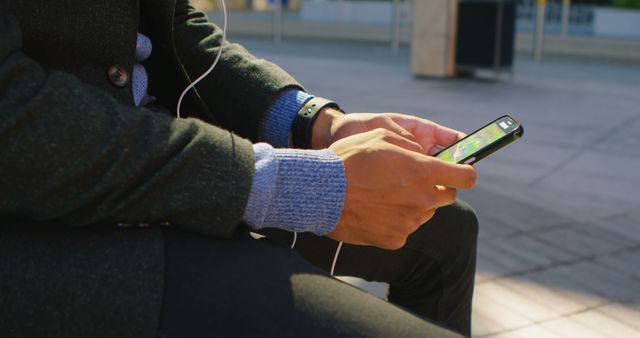 Man sitting outdoors and using smartphone while wearing earphones. Left hand resting on lap, right hand holding phone, dressed in casual outfit with a sweater and jacket. Suitable for use in contexts related to technology, mobile apps, outdoor activities, urban lifestyle, and casual wear.