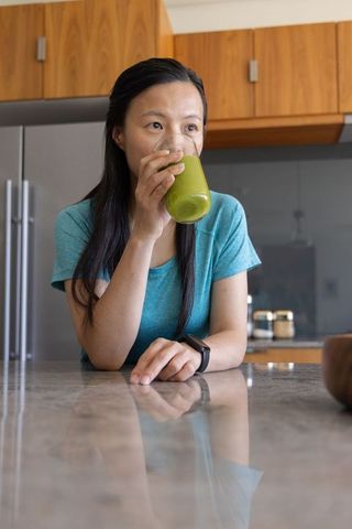 Young woman drinking green smoothie at kitchen island wearing smartwatch, wellness routine