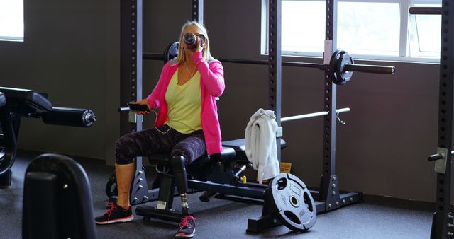 Senior woman with a prosthetic leg sitting at the gym is drinking water from a bottle after a workout. This image is useful for promoting healthy lifestyles, adaptive fitness programs, or inclusivity in sports and fitness for people with disabilities.