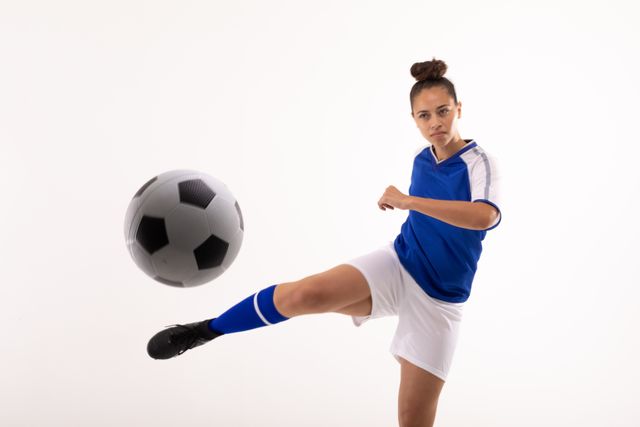 This image shows a confident young female soccer player in action, about to kick a soccer ball. She is wearing a blue and white uniform, and the background is plain white. This image can be used for sports promotions, athletic training materials, women's sports empowerment campaigns, and advertisements for soccer-related products.