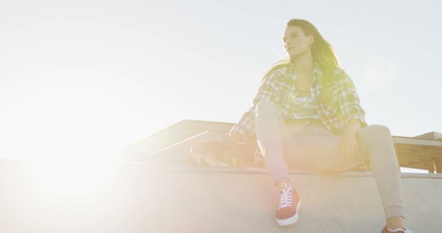 Young woman sitting on a concrete wall in an urban environment with bright sunlight. She wears casual clothing and red sneakers, exhibiting a confident and relaxed demeanor. This image is perfect for campaigns focusing on youth culture, urban fashion, or promoting outdoor activities in a city setting.
