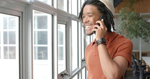 Young African American man smiling and talking on smartphone by window in coworking office