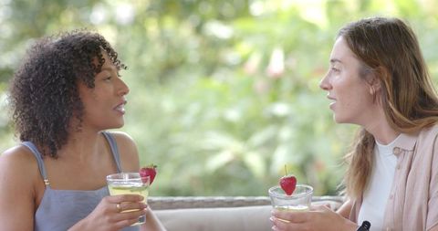 Multiracial friends enjoying strawberry cocktails on covered patio sofa, chatting