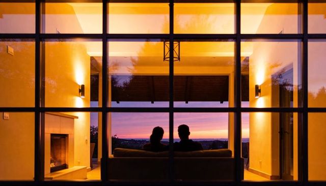 Couple watching sunset through large paned window with glowing fireplace and cozy sofa at dusk