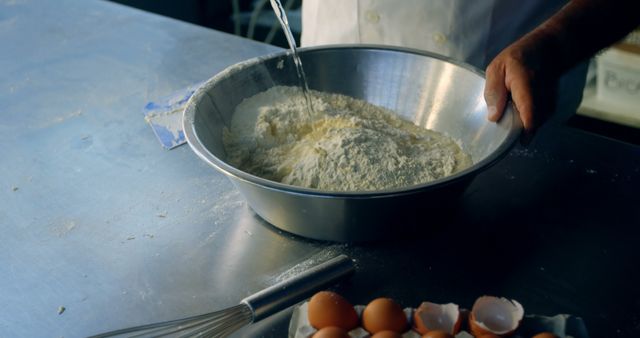Depicts process of a baker mixing flour and water in a metal bowl on a kitchen counter. Fresh eggs, cracked eggshells, and a whisk are visible, showing a baking preparation scene. Suitable for content related to culinary arts, recipes, food preparation, and bakery-related tutorials or articles.