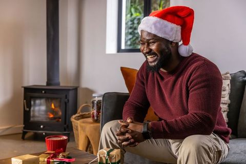 African american man wearing santa hat laughing by fireplace with wrapped christmas gifts