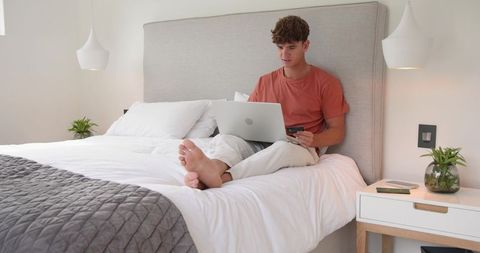 Young man shopping online and working on laptop from bed in minimalist neutral bedroom