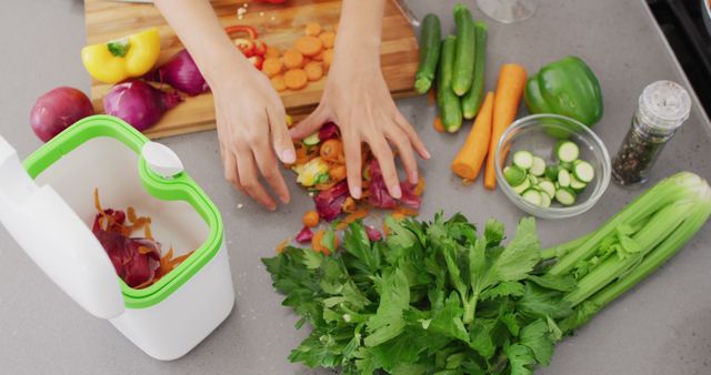 A person is preparing fresh vegetables in a modern kitchen with a compost bin nearby. Various vegetables like celery, carrots, green bell peppers, and zucchini are visible, indicating a focus on healthy cooking. This image can be used for recipes, healthy eating promotions, food blogs, or sustainability-related content.