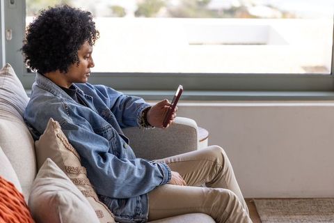 African american woman sitting on sofa holding smartphone near window in casual denim