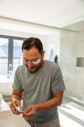 Asian man applying under-eye hydrogel patch while leaning over modern bathroom vanity