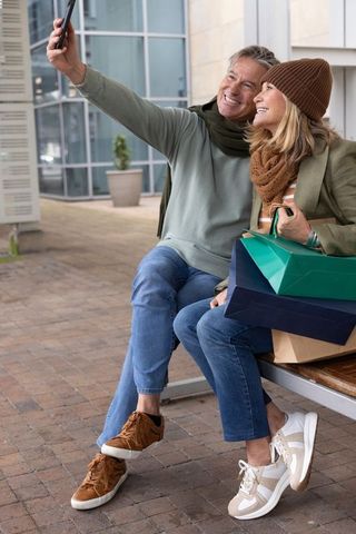 Senior couple taking selfie on urban bench during shopping trip, smiling with bags