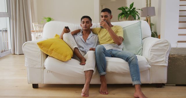 Smiling biracial gay male couple sitting on sofa embracing watching tv. staying at home in isolation during quarantine lockdown.