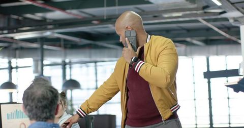 Man in Mustard Jacket Pointing at Chart While Talking on Smartphone in Open Office