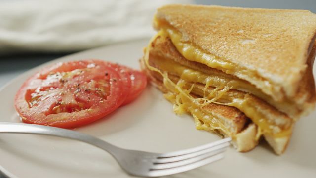 Close-up focus on grilled cheese sandwich with golden, melted cheddar cheese, served next to fresh sliced tomatoes sprinkled with black pepper on white plate. Ideal for promoting breakfast options, homemade meals, comfort food, culinary blogs, and food-related advertising. Versatile imagery for websites, menus, and social media posts.