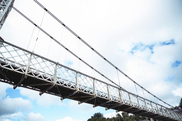 Low angle view of a historic suspension bridge with a blue sky and clouds in the background. Ideal for use in travel brochures, engineering presentations, architectural studies, and scenic photography collections.