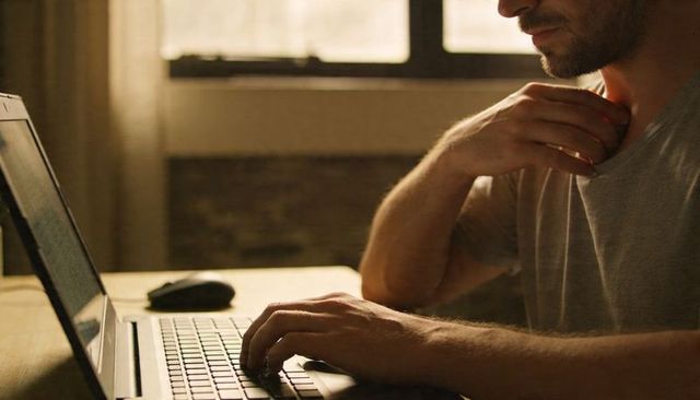 Man typing on laptop in warm daylight at wooden desk working from home focused