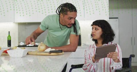 Couple collaborating in kitchen, wheelchair user holding tablet, partner offering onion