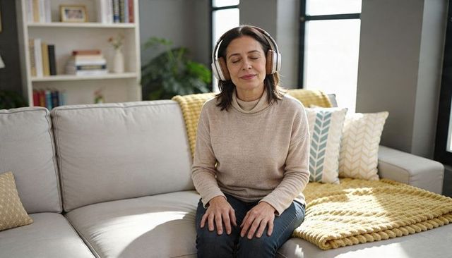 Middle-aged woman relaxing on sunlit sofa wearing headphones and cozy knit sweater
