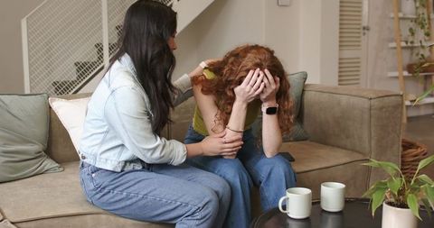 Asian woman comforting crying redhead friend on sofa, emotional support and empathy