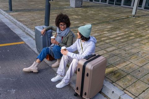 Diverse female travelers sitting on urban curb holding coffee and rolling luggage