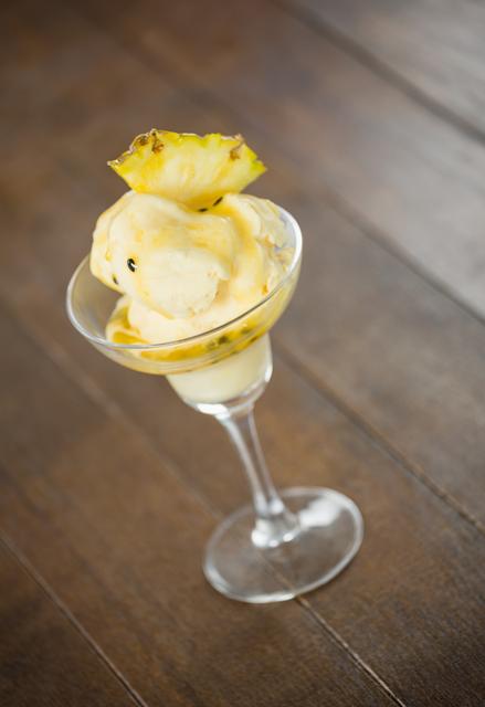 Delicious pineapple-flavored ice cream served in a clear glass, decorated with a pineapple wedge. Ideal for use in food blogs, summer menu promotions, tropical-themed events, and culinary presentations. The wooden background adds a rustic touch, enhancing the visual appeal.