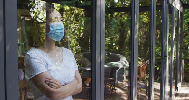 Woman standing indoors, wearing a mask, looking outside through a window with a pensive expression. Sunlight filters through trees reflecting on the glass surface. This image can be used in stories, articles, or campaigns about self-isolation, mental health, quarantine effects during a pandemic, and COVID-19 safety measures.