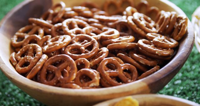 A wooden bowl filled with crunchy pretzels sits on a textured green surface, with copy space. Pretzels are a popular snack choice, often enjoyed for their salty flavor and satisfying crunch.