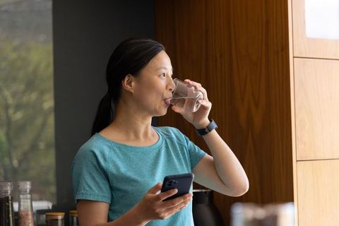 Asian woman drinking water while checking phone in sunlit modern kitchen