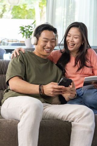Smiling asian couple relaxing on sofa sharing music and phone, listening and reading together