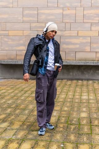 Man standing on urban plaza wearing beanie leather jacket and headphones holding coffee