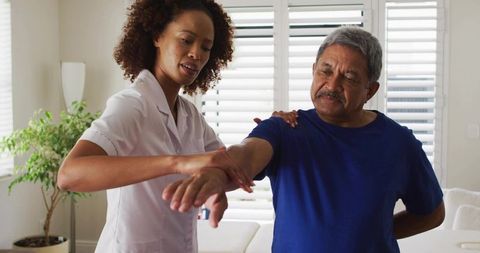 Home caregiver guiding senior man through arm mobility exercise in sunlit living room