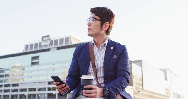Asian man holding coffee cup and smartphone while sitting on the roof of corporate park - Download Free Stock Photos Pikwizard.com