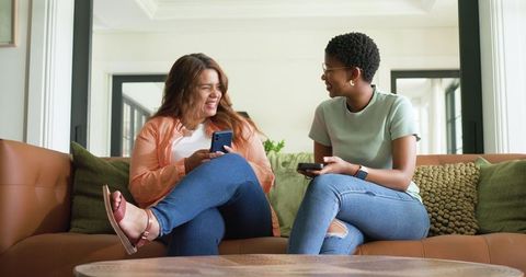 African american friends laughing on sofa holding smartphones in bright living room