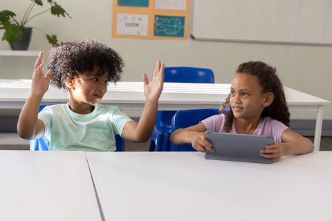 Two elementary students interacting with tablet while raising hands in bright classroom