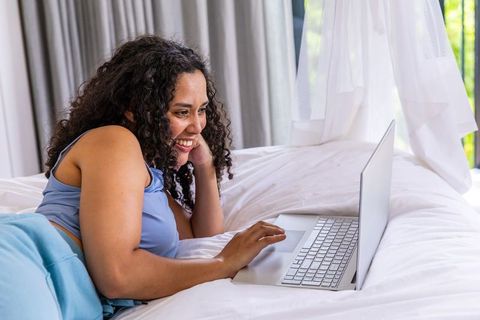 African american woman on bed using laptop, smiling while working remotely in sunlit room