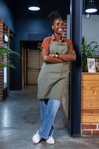Smiling african barista leaning on column in industrial cafe wearing olive apron and jeans