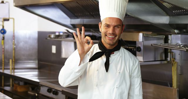 Male chef in white uniform standing in a professional kitchen, smiling and making an okay hand gesture. Ideal for use in culinary training materials, restaurant marketing, and hospitality industry promotions, highlighting professionalism and confidence in cooking.