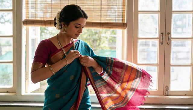 Adjusting teal silk sari pallu with gold jewelry by sunlit window, traditional south indian