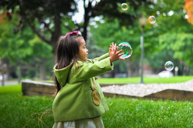 Young girl wearing green jacket playing and chasing bubbles outdoors in park. Ideal for use in materials related to childhood, happiness, outdoor activities, family life, and playful moments.