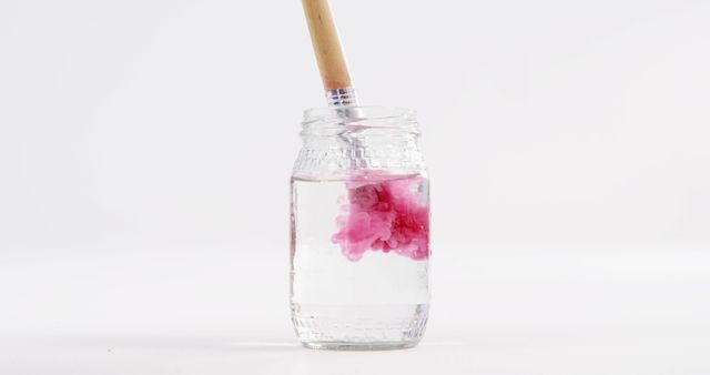 Pink paint dissolving in a glass jar of water, mixing with a wooden brush. Illustrates creative process and use of art supplies, perfect for blog posts on painting techniques, educational material for art classes, or creative product advertisements.