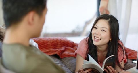 Asian couple relaxing on low sofa, woman holding book, smiling, cozy home reading moment