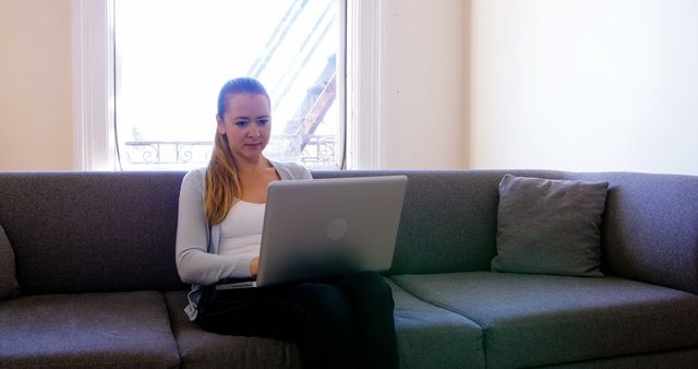 Young woman sitting on gray sofa working on laptop in bright living room with large window. Suitable for topics related to remote work, modern lifestyle, home office, online business, or casual productivity.