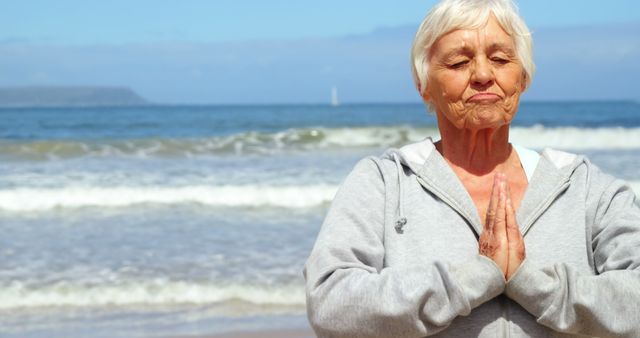 A senior Caucasian woman practices yoga on the beach, with copy space. Her peaceful expression and the ocean backdrop emphasize the tranquility and health benefits of such mindful exercises.