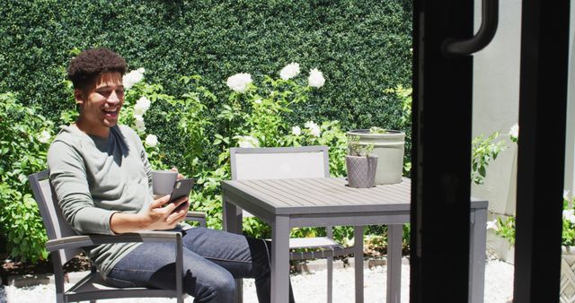 Young man sitting at patio table with smartphone and coffee, enjoying break and smiling, surrounded by green garden and white flowers. Perfect for themes of relaxation, nature, digital lifestyle, and happy moments.