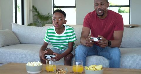 African American Father and Son Playing Video Games on Sofa with Snacks and Controllers