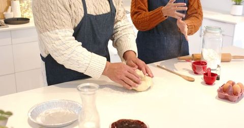 Couple kneading dough together on bright kitchen countertop wearing denim aprons